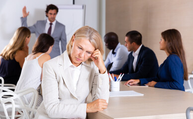 Portrait of frustrated mature woman sitting in office on background with angry boss scolding subordinates
