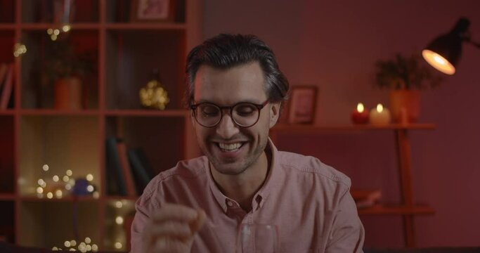 Joyful man in glasses cheering with wineglass and talking while having online conversation. Caucasian happy guy smiling and gesturing while looking to camera and sitting at home.