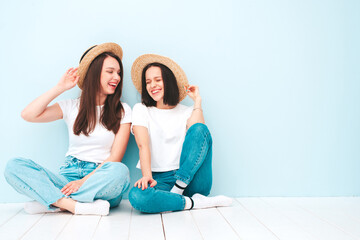 Fototapeta na wymiar Two young beautiful smiling hipster female in trendy summer white t-shirt and jeans clothes.Sexy carefree women posing near light blue wall in studio.Positive models in hats