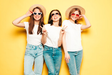 Obraz premium Three young beautiful smiling hipster female in trendy summer white t-shirt and jeans clothes.Sexy carefree women posing near yellow wall in studio.Positive models in hats show peace sign
