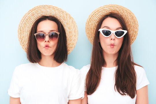 Two young beautiful smiling hipster female in trendy summer white t-shirt and jeans clothes.Sexy carefree women posing near light blue wall in studio.Positive models in hats.Making fish face