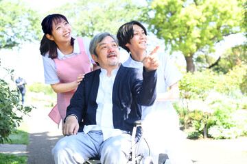 屋外で散歩をする介護ヘルパーの男女と車椅子のシニア男性 