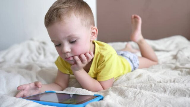 Children's technical addiction. Little preschool boy using digital tablet device lying on carpet floor alone. A small child is holding a computer surfing the Internet while playing a game at home