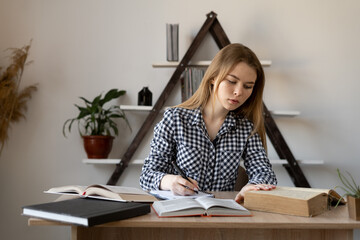 Serious focused Caucasian college girl studying at home sitting at a table with open books. European girl teacher tutor to write in a notebook teaches online remotely and prepares for a call.