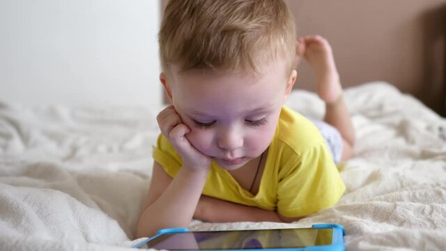 Children's technical addiction. Little preschool boy using digital tablet device lying on carpet floor alone. A small child is holding a computer surfing the Internet while playing a game at home