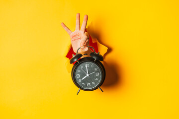 Hand shows hello gesture and black retro alarm clock out of hole in torn colored paper wall. No...