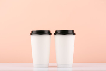 Two white disposable carton coffee or tea cups with black caps on white table against beige background with copy space. Concept of hot drinks
