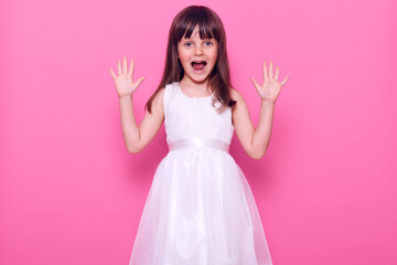 Small happy dark haired female in white dress looking directly at camera and yelling happily, celebrating positive and long-awaited event, isolated over pink background.