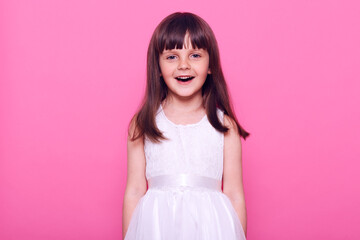 Cute smiling girl wearing white dress looking directly at camera with happy facial expression, having dark hair, positive mood, isolated over pink background.