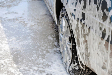 The rear wheel of the car and part of the body are covered with detergent foam. Advertising of cleaning services at a car wash with space for text.