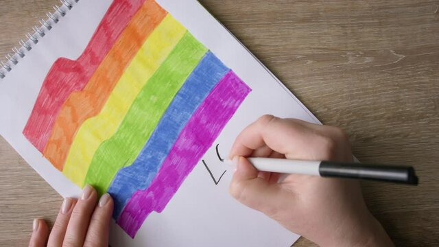 Female hands write down LOVE IS LOVE on drawing with rainbow colors flag.