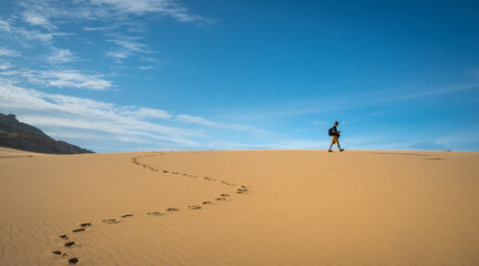Fototapeta na wymiar Traveler cum hiker walking in the desert leaving trail of footprints. Bright blue sky at the horizon.