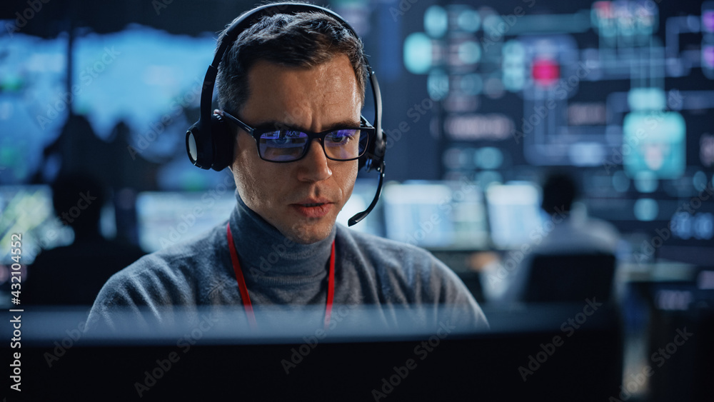 Poster Portrait of Professional IT Technical Support Specialist Working on Computer in Monitoring Control Room with Digital Screens. Employee Wears Headphones with Mic and Talking on a Call.