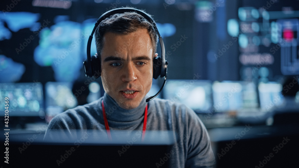 Poster Portrait of Professional IT Technical Support Specialist Working on Computer in Monitoring Control Room with Digital Screens. Employee Wears Headphones with Mic and Talking on a Call.