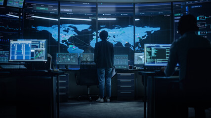 Diverse Multiethnic IT Technical Support Manager with Curved Hair Stands next to Workplace in Monitoring Control Room Office with Big Digital Screens with Server Technical Blockchain Data.