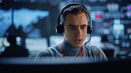 Portrait of Professional IT Technical Support Specialist Working on Computer in Monitoring Control Room with Digital Screens. Employee Wears Headphones with Mic and Talking on a Call.