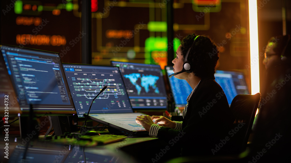 Poster Multiethnic Female IT Technical Support Specialist Talking with a Client in Headphones and Working on Desktop Computer in Monitoring Control Room with Big Digital Screens with Server Blockchain Data.