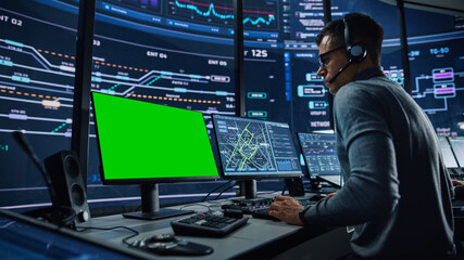Professional IT Technical Support Specialist and Software Developer Working on Computer with Green Screen Mock Up Display in Monitoring Control Room with Digital Screens. Employee Wears Headphones.