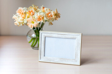 Portrait white picture frame mockup on wooden table. Modern glass vase with Daffodils. White wall background. Scandinavian interior. 