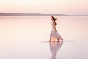 Young blonde woman in an evening airy pastel pink, powdery dress stands barefoot on white crystallized salt. Girl with natural make-up, hair is developing. Salt mining trip, walking on water at sunset