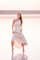 Young blonde woman in an evening airy pastel pink, powdery dress stands barefoot on white crystallized salt. Girl with natural make-up, hair is developing. Salt mining trip, walking on water at sunset