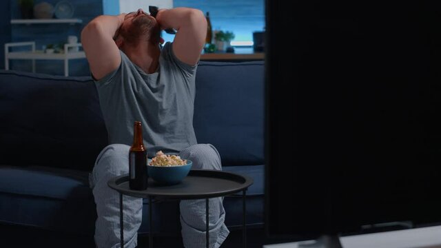 Upset man football supporter watching sport game supporting favourite team eating nervos popcorn after losing championship. Dissapointed male gesturing talking alone and drinking beer