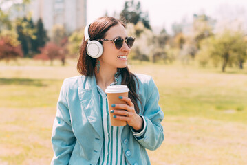 A cheerful young woman in park is drinking her hot drink and looking away is listening to the music in her headphones