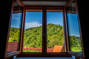 Wide open window through which you can see the green landscape of nature with a wooded mountain peak and the blue sky on a sunny summer day. Natural ventilation of the house or room.