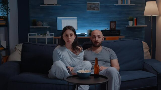 Shocked astonished young couple looking at documentary show at tv having surprised facial expression, eating popcorn sitting on sofa. Concentrated adults watching tv late night enjoying free time