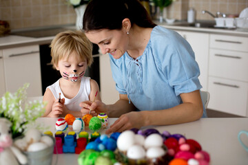 Beautiful blond child, toddler boy, painting easter eggs with mother at home, making easter wreath