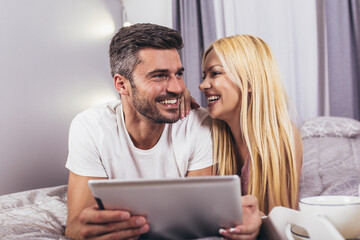 Smiling relaxed young couple using digital tablet in bed at home