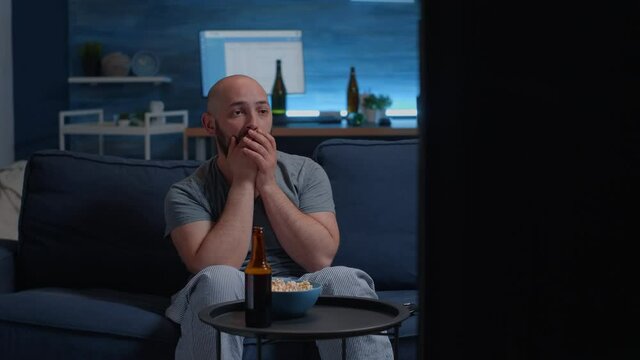 Emotional young guy watching disgusting tv program movie alone in living room, feeling confused shocked about horror film, violence in social media, awful news or morbid shows. Man enjoying free time