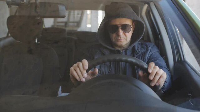 A man in black glasses and a hoodie at the wheel of a car is secretly shooting on a long-focus lens.