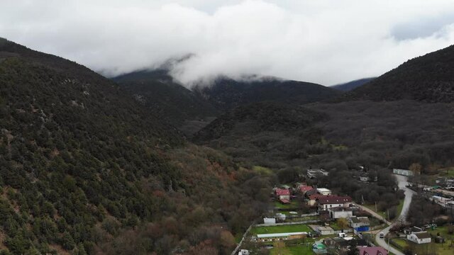 Small Town At The Foot Of The Alps. Stock Footage. Aerial View From A Drone To The Tops Of Mountains In White Clouds. Natural Present Background. A Truck Drives By Far Below, Personifying The Current