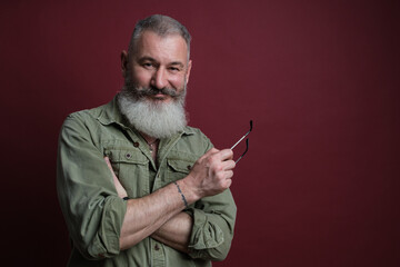 Mature bearded caucasian man looks slyly at camera with eyeglasses in hand, red background