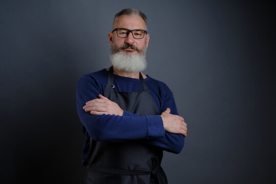 Portrait of handsome mature bearded man dressed leather apron isolated on black background, caucasian workman with beard smiling