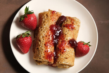 Pancakes with strawberry jam on plate. Pancakeshin rolls poured berry jam and decorated fresh strawberries on brown background. Top view. 