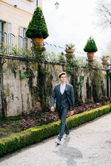 Serious groom walks through a green park against the backdrop of a stone fence and old buildings. Lake Como