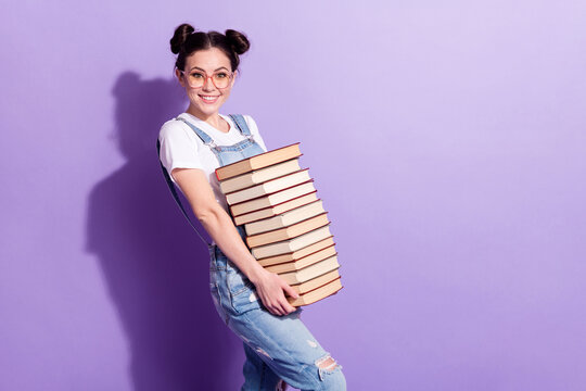 Photo of funny pretty young woman dressed denim overall glasses holding books stack empty space isolated purple color background