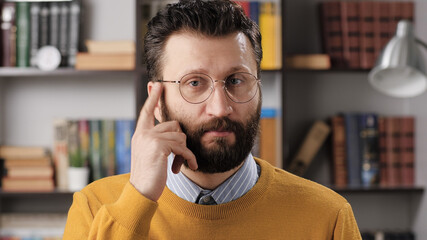 Naklejka premium Just think gesture emotion. Positive bearded man teacher or businessman with glasses looking at camera and tapping his finger on his temple, suggests thinking well