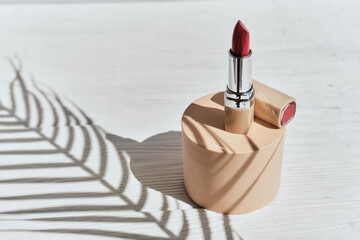 Red lipstick on a beige cylinder podium with shadows from branches of palm