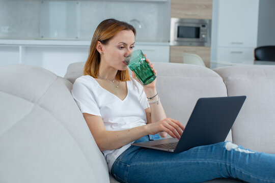 A Girl In A White T-shirt Drinks Water From A Glass, Works Behind A Laptop Sitting At Home On The Couch. High Quality Photo