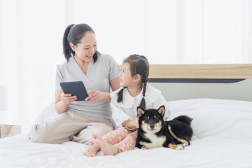 Happy time together. Young mother and small daughter playing with Shiba inu dog on bed. Girls and mother and pet dog using tablet at home together happy.