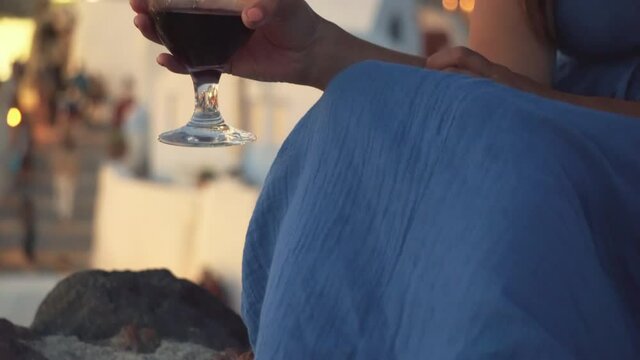 Partial view of woman holding homemade sweet rose wine in small shot glass sitting on Oia castle and view of Greek island Santorini sunset.Feet of elegant woman sitting on castle wall in Oia, Santorin