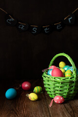 A green basket with Easter eggs stands on a wooden table, on a dark background, the word Easter in the dark, still life, vertical photo