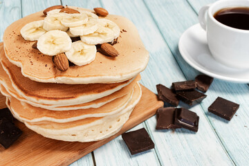 Homemade pancakes with banana and chocolate on a light blue wooden background with a cup of coffee in a close-up