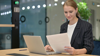 Serious Young Businesswoman with Laptop Reading Documents in Office 