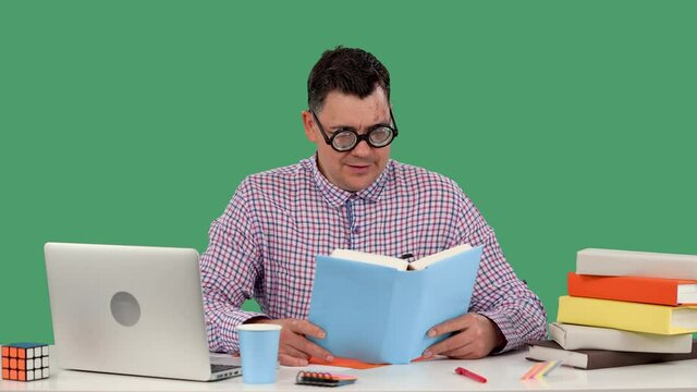 A man in glasses with with thick lenses sits at a desk in front of a laptop and reads a book, lifts his glasses and says. Portrait of a man in the studio on a green screen. Slow motion. Close up.