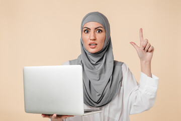 Smart young Middle Eastern muslim islamic arabian student female woman using laptop for online learning , webinars having idea isolated over beige background. office worker girl in hijab using laptop