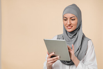  islamic muslim arabian Middle Eastern young woman student using digital tablet isolated over beige background. Clever young female office worker in grey hijab chatting on tablet, sending messages.